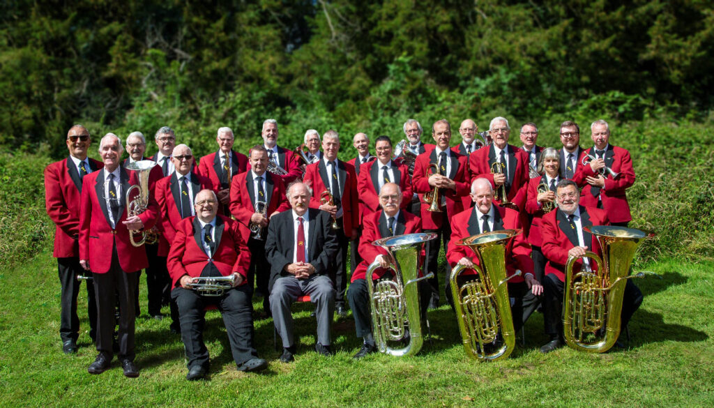 Keynsham Brass Band in red jackets and black trousers posed outdoors with tubas, trumpets and other brass instruments on a grass lawn in Henleaze, Bristol.