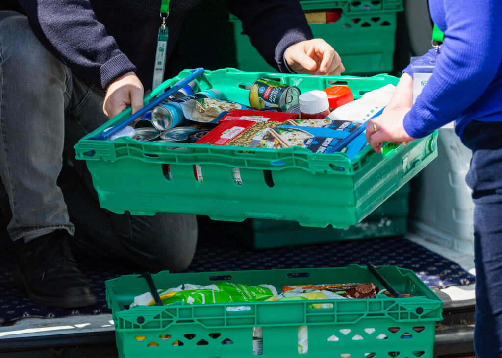 Two people sort food items into green plastic crates inside a van. One holds a container filled with cans and bottles.