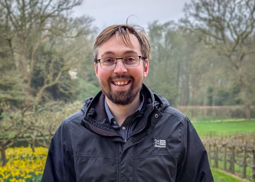 A man wearing glasses and a dark jacket with a Karrimor logo stands in a garden with trees and yellow daffodils.