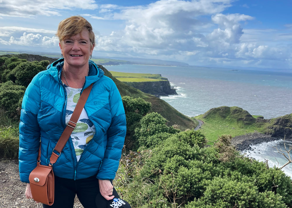 A woman in a teal puffer jacket and dark trousers stands on a rocky overlook overlooking the ocean. She carries a brown leather bag and is smiling at the camera.