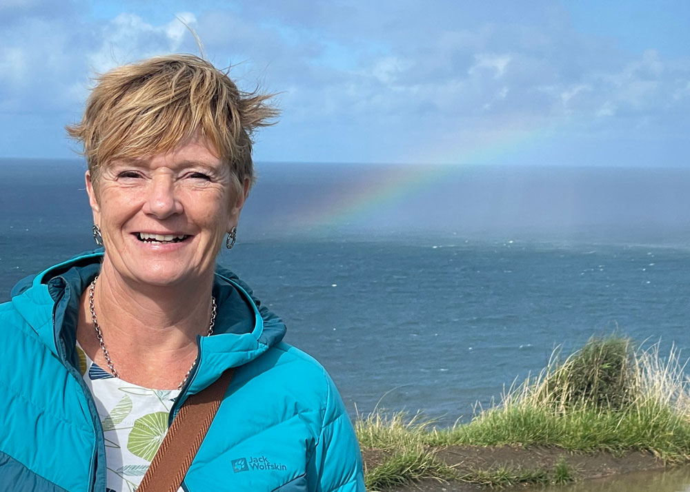 A woman in a teal jacket stands outdoors near a grassy bank overlooking the sea. A rainbow arches across the sky behind her.