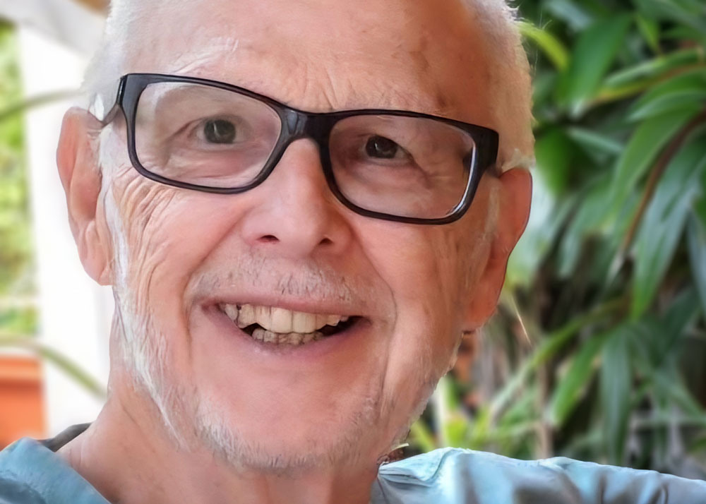 An older man with glasses smiles at the camera. He wears a light blue shirt. Green foliage is visible in the blurred