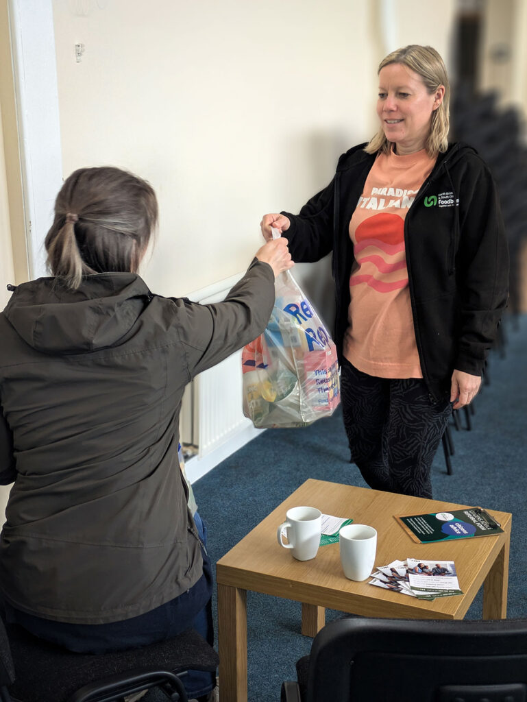 A woman hands a plastic bag containing food items to a person seated at a table with mugs and leaflets.