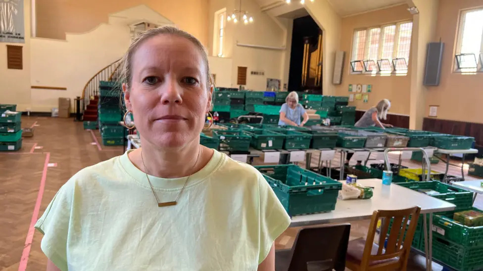 ellen wood warehouse Ellen Wood stands in the food bank warehouse, wearing a pale green t-shirt and a necklace, amidst stacks of green plastic crates.